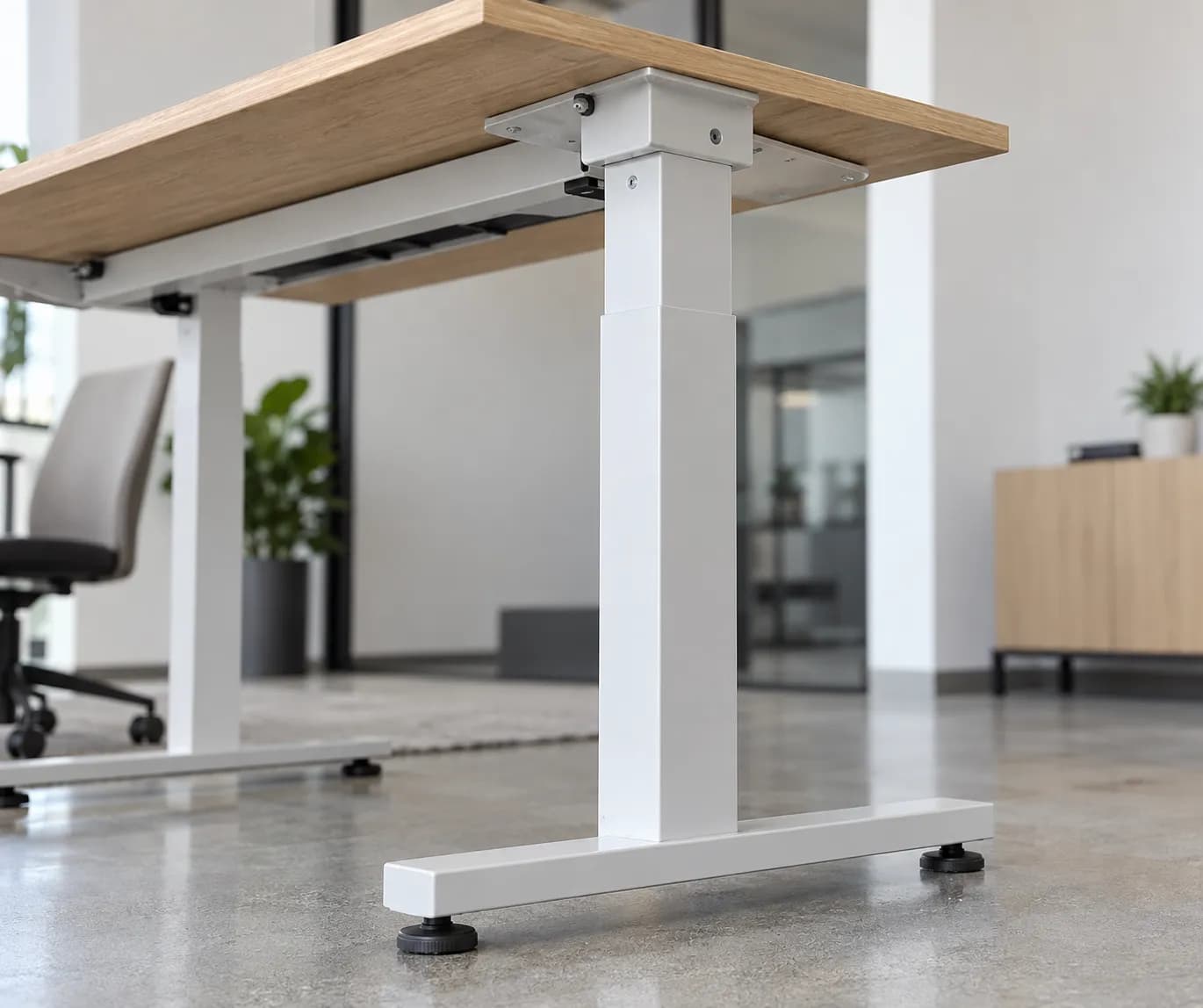 Close view of an unbranded height-adjustable desk frame, tabletop edge and leveling foot at a commercial workspace floor